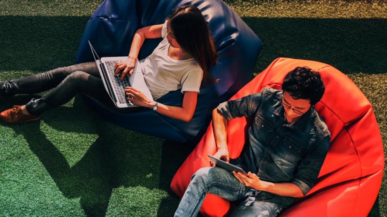 Young Asian couple using laptop notebook and digital tablet together in modern public park, sit on bean bag, top view with copy space on grass.