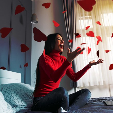 Beautiful, happy, young woman throwing red heart-shapes in the air, sitting on the bed.