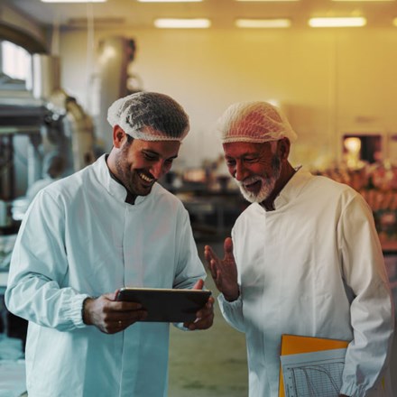 Two cheerful male colleagues in sterile clothes standing in food factory and looking at sales documents on digital tablet.
