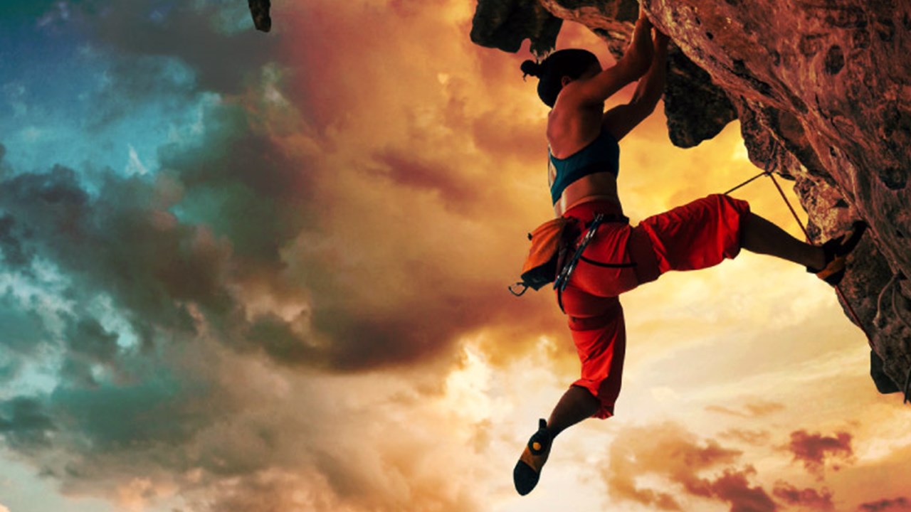 Athletic woman climbing on overhanging cliff rock with sunset sky background.