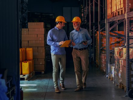 Two business partners in formal wear and with protective yellow helmets on heads walking and talking about business. Younger one holding folder with data while older one using tablet.